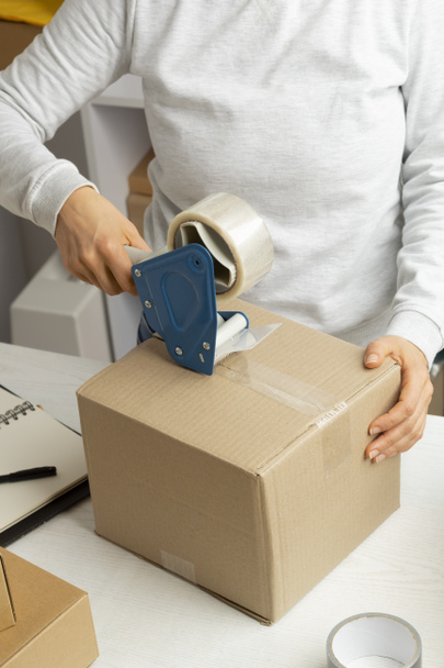 A warehouse worker packs a box of goods for delivery using an adhesive tape dispenser. - 写真・画像