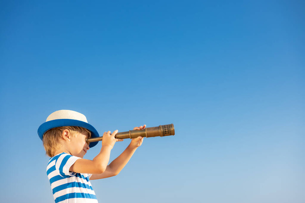 Child looking through spyglass against blue sky. Kid having fun outdoor in summer. Adventure and travel concept - Фото, изображение