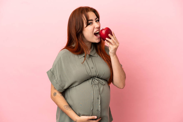 Νεαρή κοκκινομάλλα καυκάσια γυναίκα απομονωμένη σε ροζ φόντο έγκυος και κρατώντας ένα μήλο και τρώγοντας το - Φωτογραφία, εικόνα