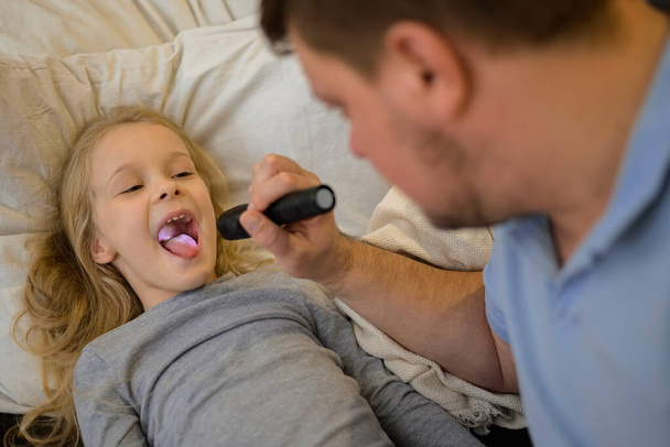 dad checks the throat of his sick child, shines a flashlight so that it is better to see. The father suspects that his daughter is ill. The child is not feeling well. the concept of equality in the - Photo, image