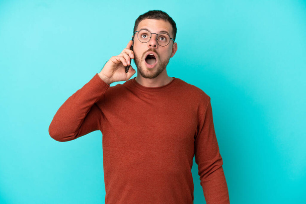 Young Brazilian man using mobile phone isolated on blue background looking up and with surprised expression - Foto, Imagem