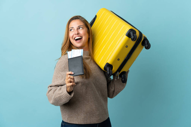 Traveler middle age woman holding a suitcase isolated on blue background in vacation with suitcase and passport - 写真・画像
