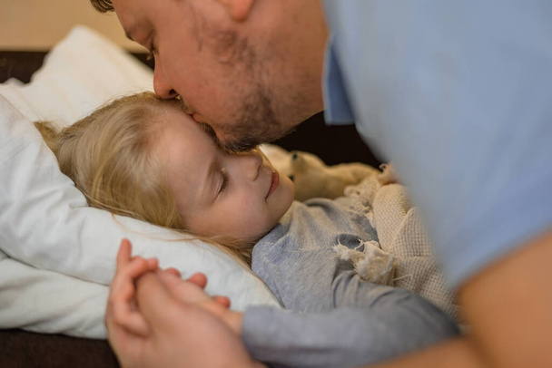 dad kisses his sick child on the forehead before bed. Father with tenderness and care kisses his daughter. the concept of equality in the family between father and mother. Father on sick leave with - Photo, Image