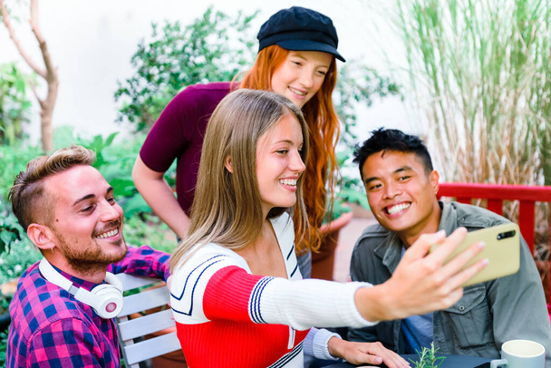 Monikulttuurinen ryhmä onnellisia nuoria ystäviä ottamassa selfietä yhdessä matkapuhelimella nauraen ja hymyillen, kun he poseeraavat ryhmänä ulkona - Valokuva, kuva