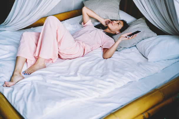 Millennial hipster girl in sleepwear lying at homey bead and listening audio book during Sunday morning in comfortable flat, Asian woman in electronic earphones enjoying smartphone music playlist - Valokuva, kuva