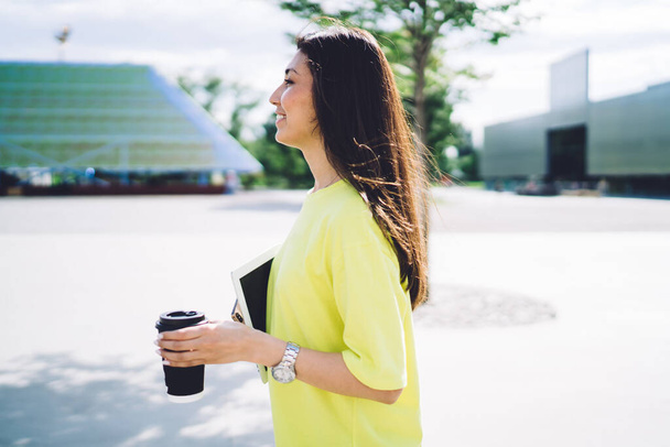 Smiling hipster girl walking on urban setting during spring sunny day holding coffee to go cup, positive woman in casual wear strolling during free time with takeaway enjoying rest on city street - Фото, изображение