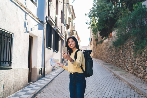 Positive ethnic female in casual clothes and backpack standing on street and talking on video chat on smartphone while smiling and holding map - Fotoğraf, Görsel
