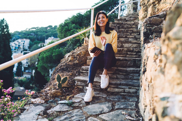 Full body of cheerful Asian female in casual clothes with toothy smile sitting on stone stairs and reading book during walk - 写真・画像