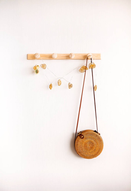 Minimalistic image of round rattan boho crossbody bag hanging on wooden hanger against white wall. Fashion and bohemian style accessories concept. Minimal home interior design - 写真・画像