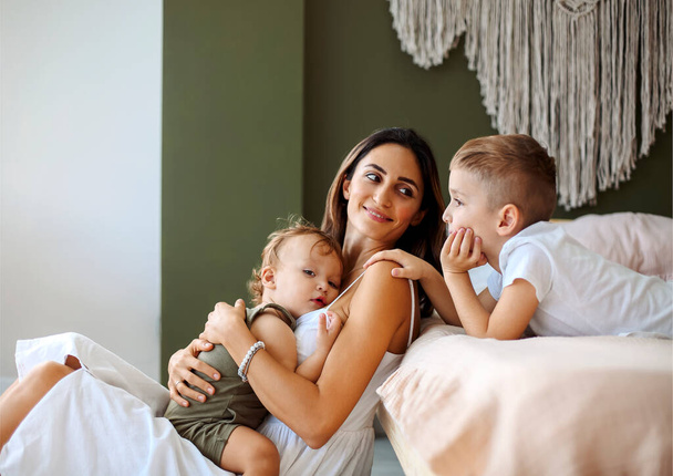 Молода сім'я красива мати з двома синами різного віку проводить час вдома, любляча мама тримає дитину в руках і піднімає його спати з допомогою і підтримкою старшої дитини
 - Фото, зображення