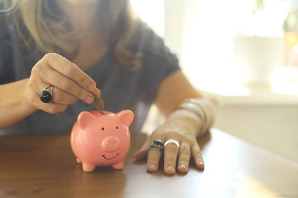 Saving money. Cropped shot of woman putting coin in ceramic or porcelain piggy bank while sitting at table at home, selective focus. Basics of budgeting, thrift and savings concept - Photo, Image