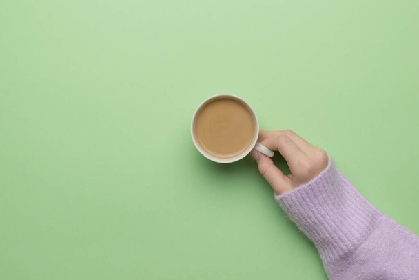 Beautiful hand of a girl in a warm sweater with a cup of coffee on a green background. The concept of drinking a popular drink. Flat lay. - Фото, зображення