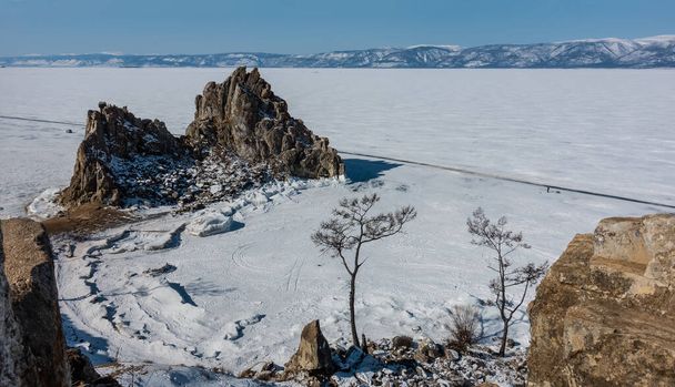 A picturesque granite double-headed rock rises above a frozen lake. On the snow-covered ice, you can see a highway, a car, a lonely tiny silhouette of a man. Bare trees in the foreground. Baikal. - 写真・画像