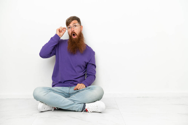 Joven hombre caucásico rojizo sentado en el suelo aislado sobre fondo blanco con gafas y sorprendido - Foto, Imagen
