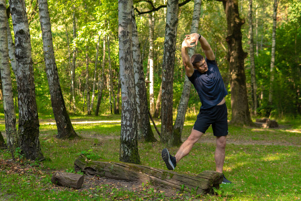 A young enduring athletic athlete is doing stretching in the forest outdoors, around the forest, oak trees.active runner workout jogger wellness motion, park outside. Adult running, feet stretches - Foto, afbeelding