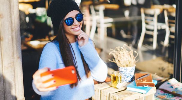 Happy millennial influencer shooting positive video vlog using smartphone front camera during leisure in sidewalk cafe, cheerful hipster girl in trendy sunglasses taking selfie pictures on mobile - Fotoğraf, Görsel