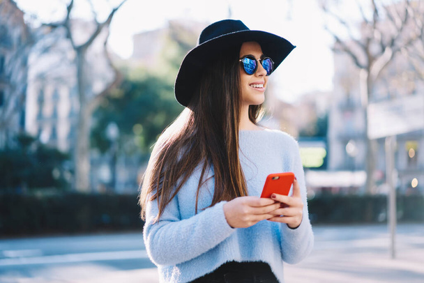 Trendy dressed female blogger with mobile technology smiling during leisure on vacations, happy Caucasian tourist enjoying getaway journey and time for media networking via smartphone gadget - Fotografie, Obrázek