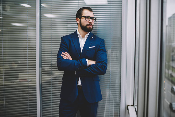Serious handsome man in elegant suit and eyeglasses with beard standing near glass wall with arms crossed and looking away in office hall - Photo, Image