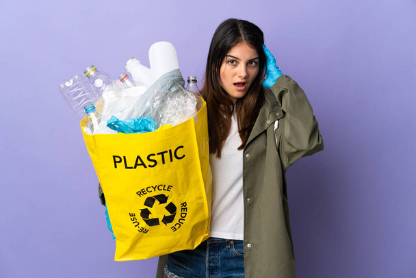 Jonge vrouw met een zak vol plastic flessen te recyclen geïsoleerd op paarse achtergrond met verrassende expressie - Foto, afbeelding