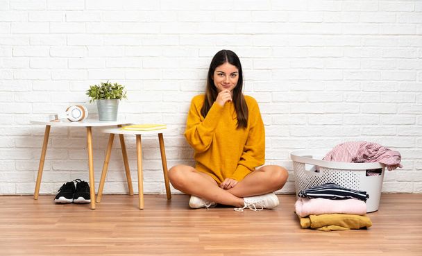 Young woman sitting on the floor at indoors with clothes basket thinking - Photo, Image