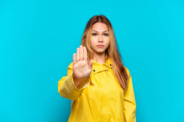 Teenager Mädchen in einem regendichten Mantel über isoliertem blauen Hintergrund macht Stop-Geste - Foto, Bild