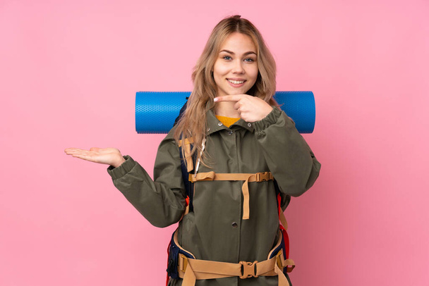 Έφηβη Ρωσίδα ορειβάτισσα με ένα μεγάλο σακίδιο, απομονωμένο σε ροζ φόντο, που κρατάει το copyspace φανταστικό στην παλάμη για να βάλει αγγελία. - Φωτογραφία, εικόνα