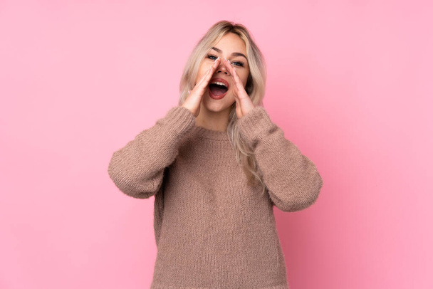tiener blond meisje het dragen van een trui over geïsoleerde roze achtergrond schreeuwen en aankondigen iets - Foto, afbeelding