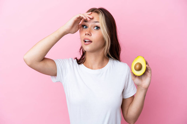Young Russian woman holding avocado isolated on pink background doing surprise gesture while looking to the side - Foto, Bild