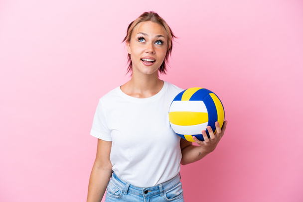 Young Russian woman playing volleyball isolated on pink background looking up and with surprised expression - Фото, изображение