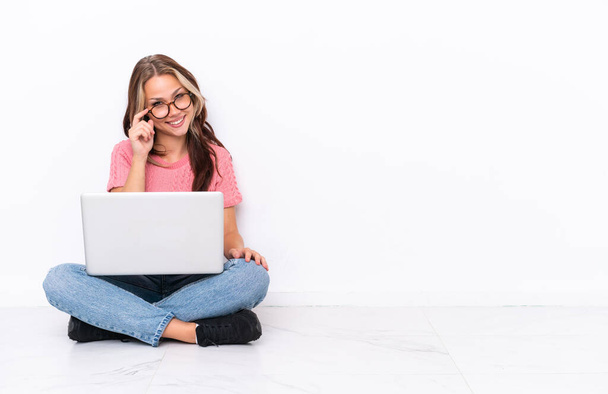 Joven chica rusa con un ordenador portátil sentado en el suelo aislado sobre fondo blanco con gafas y feliz - Foto, Imagen