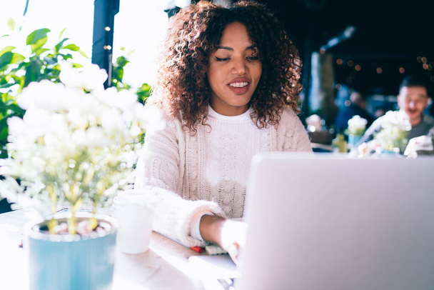 Cheerful African American female in casual clothes sitting in modern cafe and smiling while browsing laptop and working on remote project - Foto, Bild