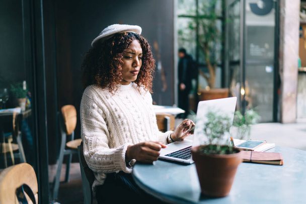 Ethnic woman freelancer with curly hair and in casual clothes sitting at table and using laptop to work on project in cozy cafe - Фото, зображення