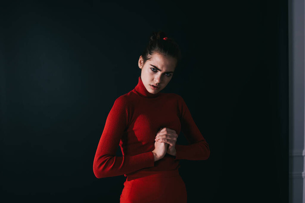 Serious woman in red outfit frowning and looking at camera while clenching fist in front of chest in dark room - Fotoğraf, Görsel