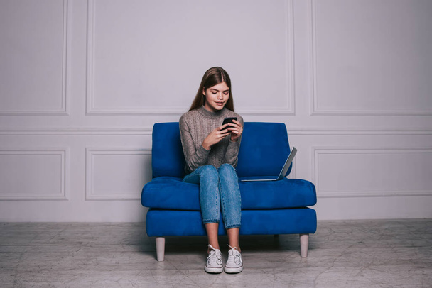Happy female student in casual outfit texting message on mobile phone after work while sitting on sofa in light room - Valokuva, kuva