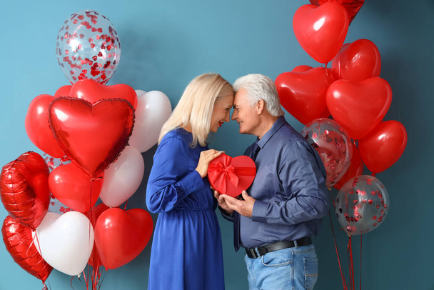 Glückliches reifes Paar mit Geschenk und Luftballons auf farbigem Hintergrund. Valentinstag-Feier - Foto, Bild