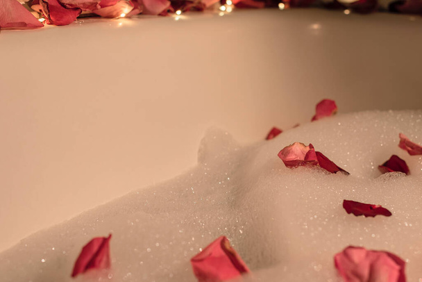 Bathtub with foam, pink rose petals and lights. Enjoying life, luxury, love concept. Valentine or other holiday greeting card with copy space - Photo, image