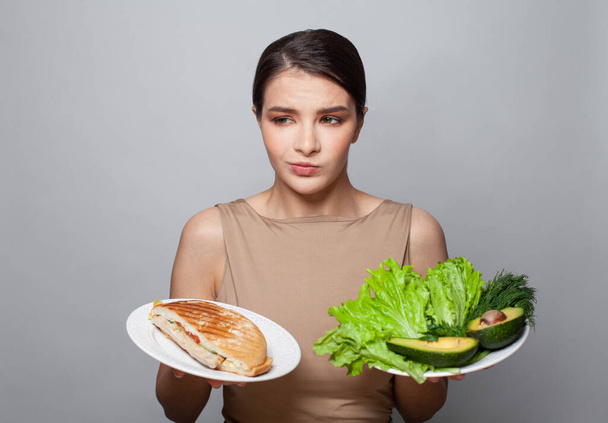 Studio shot of a young brunette woman choosing between healthy and unhealthy foods against a grey background. Woman diet choice - Photo, image