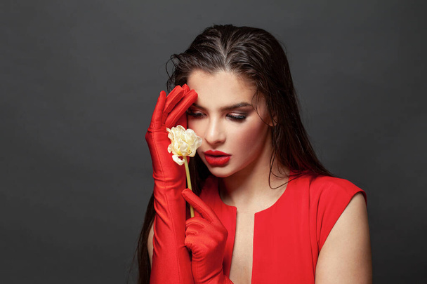 Sensual woman brunette with makeup wearing red dress and red silky gloves holding white flower on black background - Fotó, kép