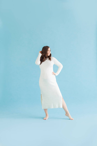 Pretty pregnant woman in white cotton dress standing on blue background - Φωτογραφία, εικόνα