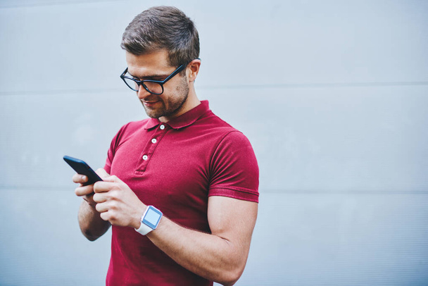 Concentrated bearded male in casual outfit browsing mobile phone while standing on street against gray wall of building in daytime - Foto, Imagem