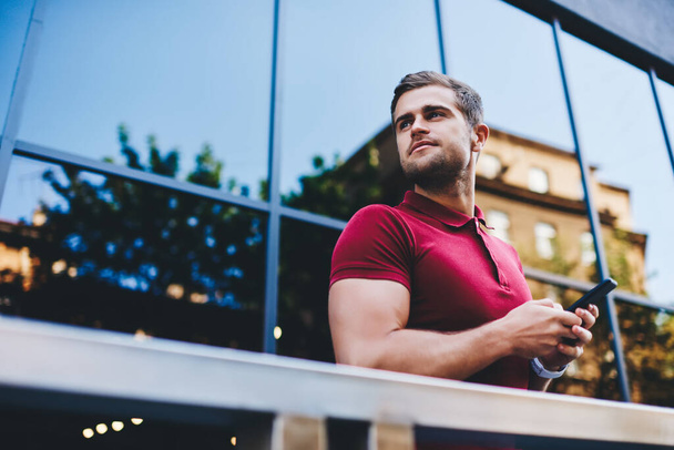 From below of pensive young hipster in polo shirt browsing smartphone and looking away beside modern office building on blurred background - Foto, imagen