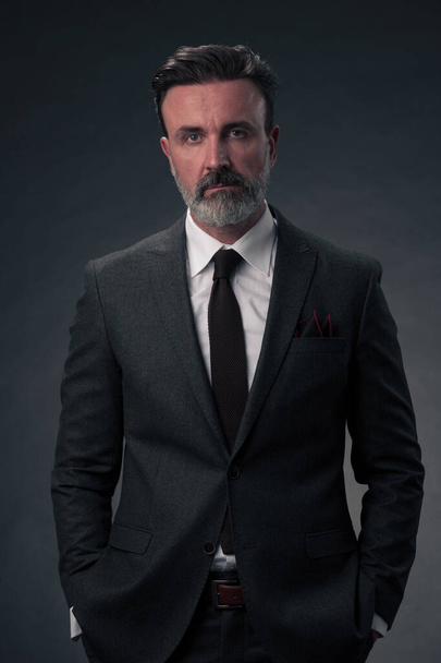 Portrait of a stylish elegant senior businessman with a beard and casual business clothes in photo studio isolated on dark background gesturing with hands. High quality photo - Foto, Bild
