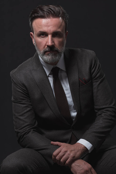 Portrait of adult businessman wearing trendy suit and sitting in modern studio on stylish chair against the black background. Horizontal mockup. High quality photo - Φωτογραφία, εικόνα