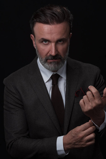 Portrait of a stylish elegant senior businessman with a beard and casual business clothes in photo studio isolated on dark background adjusting the suit - 写真・画像