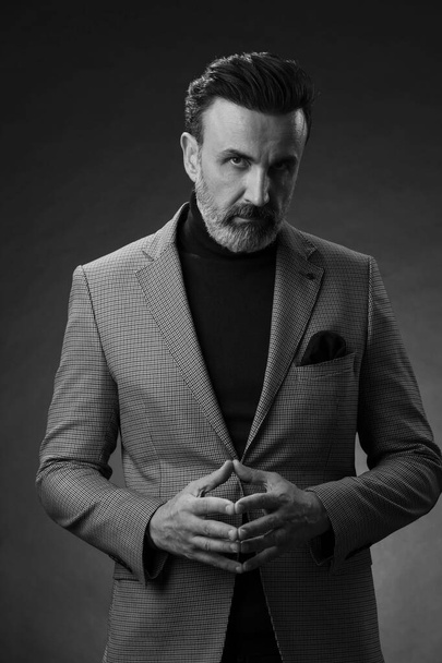 Portrait of a stylish elegant senior businessman with a beard and casual business clothes in photo studio isolated on dark background gesturing with hands. High quality photo - Фото, изображение