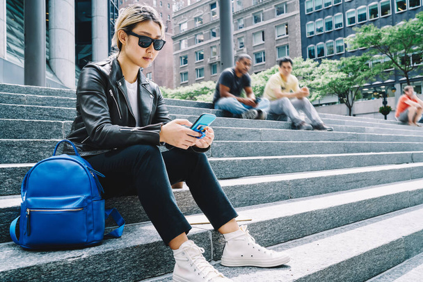 Asian hipster girl in trendy sunglasses jacket using mobile device for reading travel publication in social media, millennial tourist connecting to 4g internet for browsing web text via smartphone - Foto, Imagen
