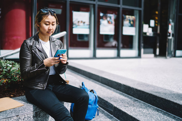 Asian hipster girl in trendy leather jacket using cellphone device for reading travel publication in social media, millennial tourist connecting to 4g internet for browsing web text via smartphone - 写真・画像
