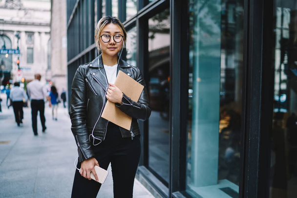 Half length portrait of millennial blogger in eyeglasses holding education sketchbook looking at camera while listening music podcast, Chinese female in electronic headphones using cellphone app - Foto, Bild