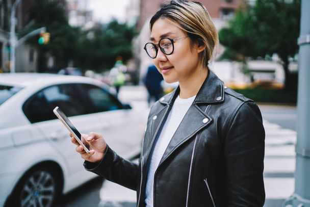 Attractive millennial blogger in classic eyeglasses installing media application while making online booking via cellphone device, Chinese hipster girl connecting to 4g internet for networking - Foto, Imagen