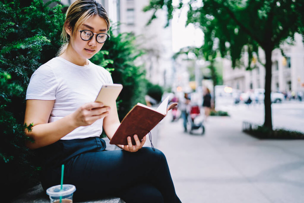 Millennial hipster girl in trendy glasses checking content text for sharing publication to social media, Chinese woman with textbook for planning spending time for smartphone networking in city - Foto, afbeelding